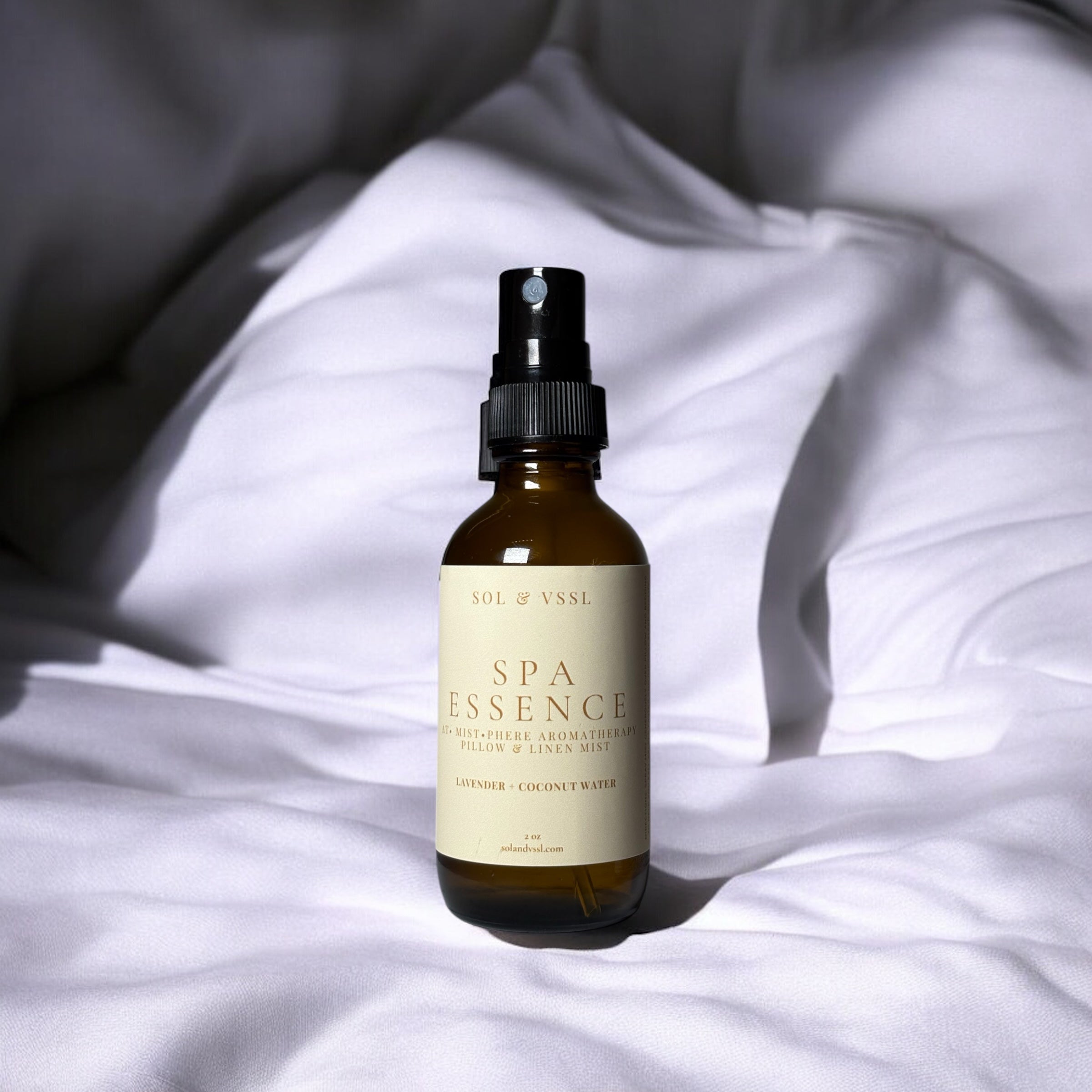 A brown amber glass spray bottle labeled 'SPA ESSENCE' with a black nozzle, placed on a bed with white sheets.
