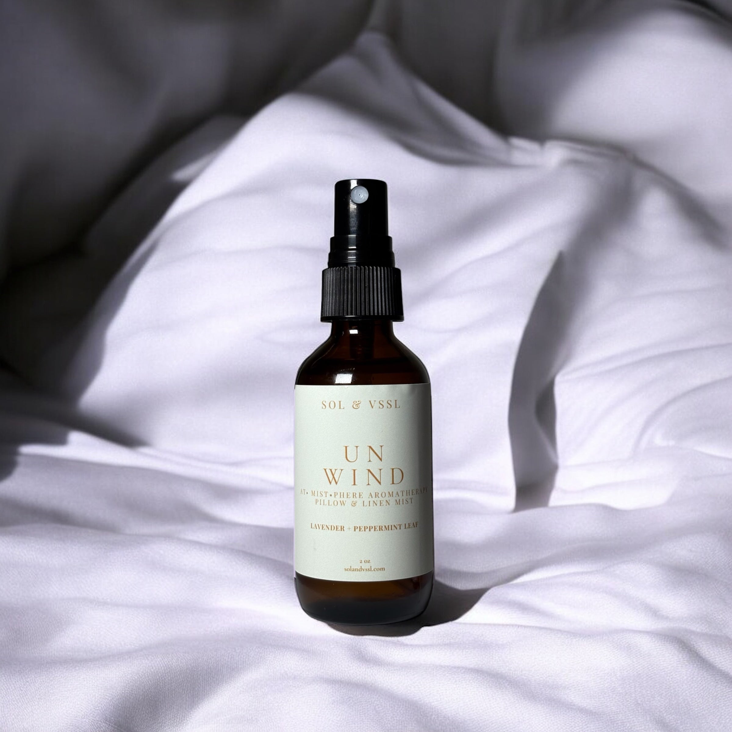 A heavy amber glass spray bottle with a black nozzle labeled 'UNWIND' placed on a bed of sheets, suggesting a calming effect for sleep.